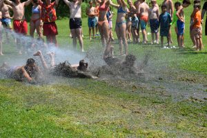 3 male campers on their stomachs sliding down a mud slide with campers and counselors cheering in the background and watching them.