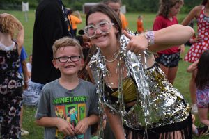 female counselor with silver glasses and silver streamers hanging off of her giving a thumbs up sign standing next to a little boy smiling.