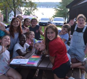 group of female campers around a picnic table smiling and waving at the camera.